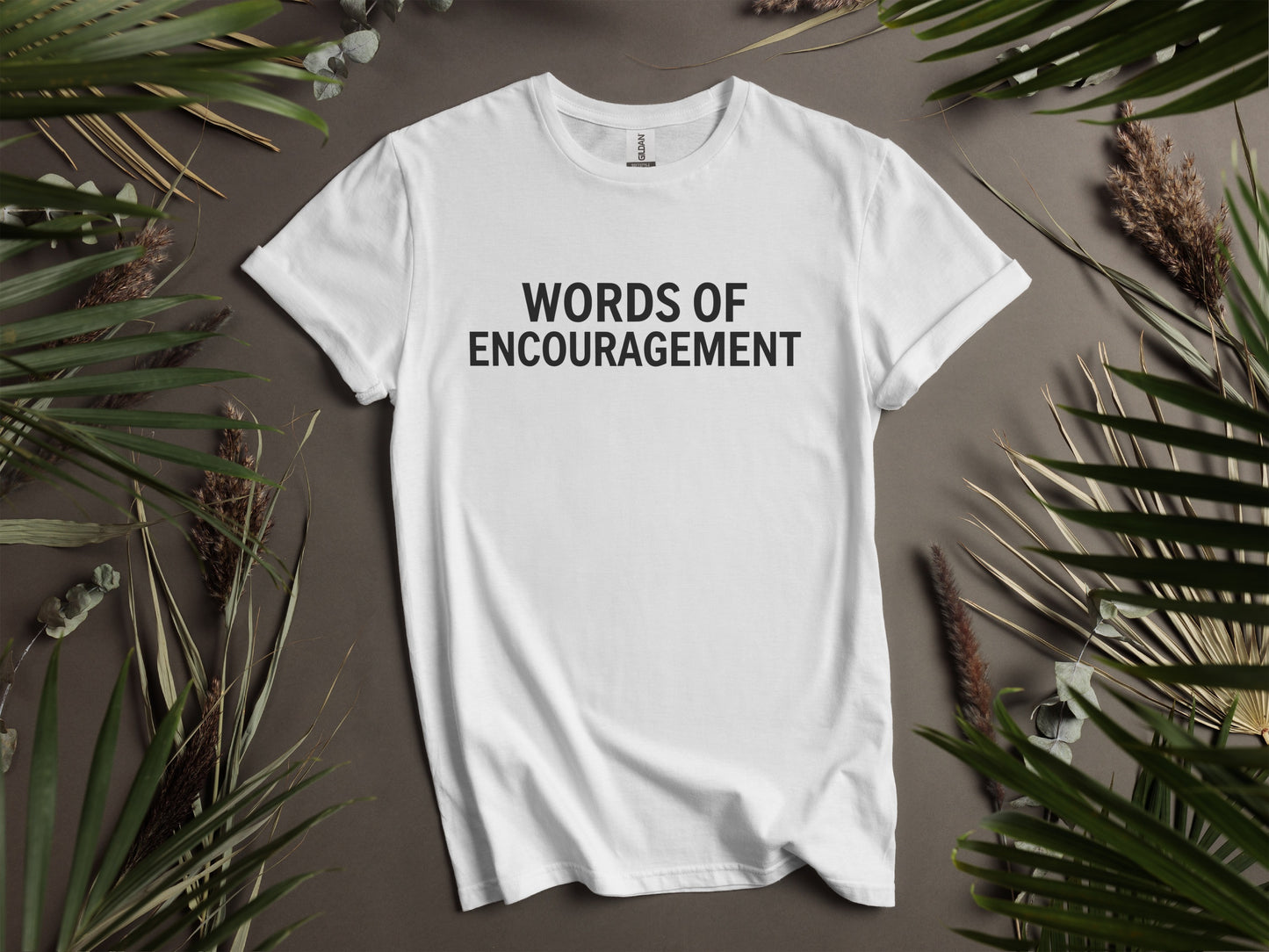 White t-shirt with "WORDS OF ENCOURAGEMENT" text, framed by dried plants.
